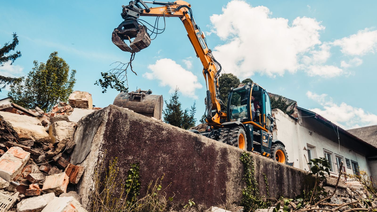 Wheeled excavator moving brick rubble beside a partly demolished building