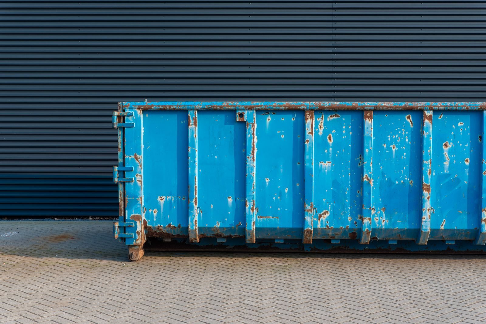 Blue roll-off dumpster parked against a dark corrugated metal wall